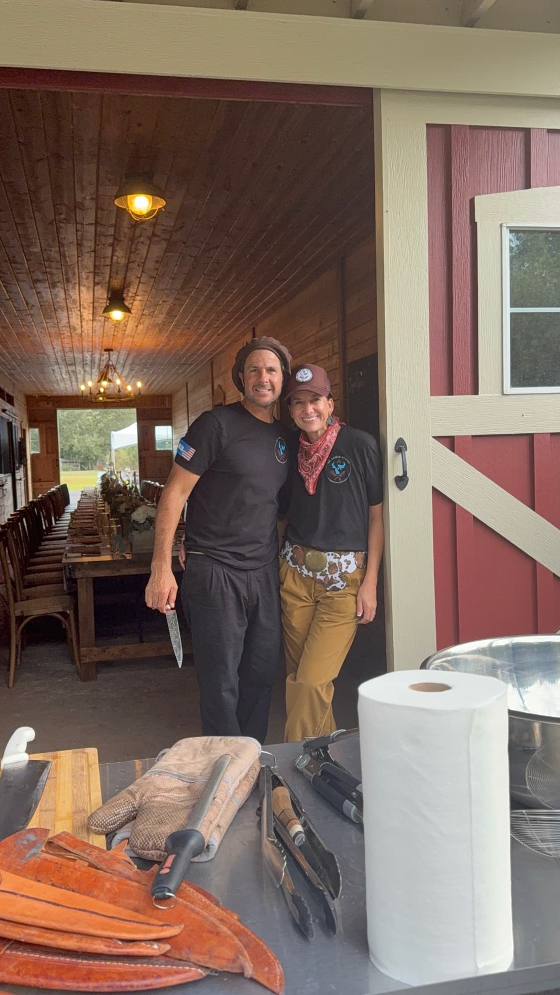 Marcos and Mandi Villanueva standing together in front of the red barn, a long table set behind them