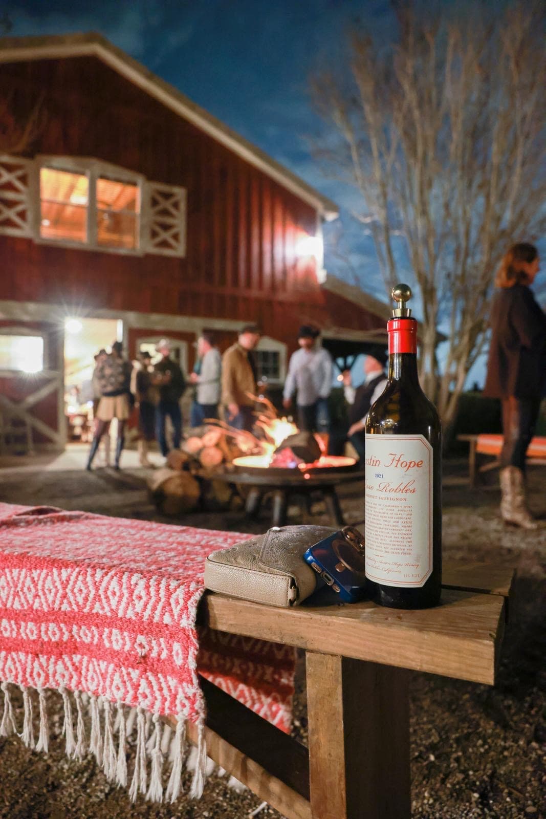 Guests gathered around a fire pit at dusk beside the glowing red barn, a bottle of wine resting on a bench